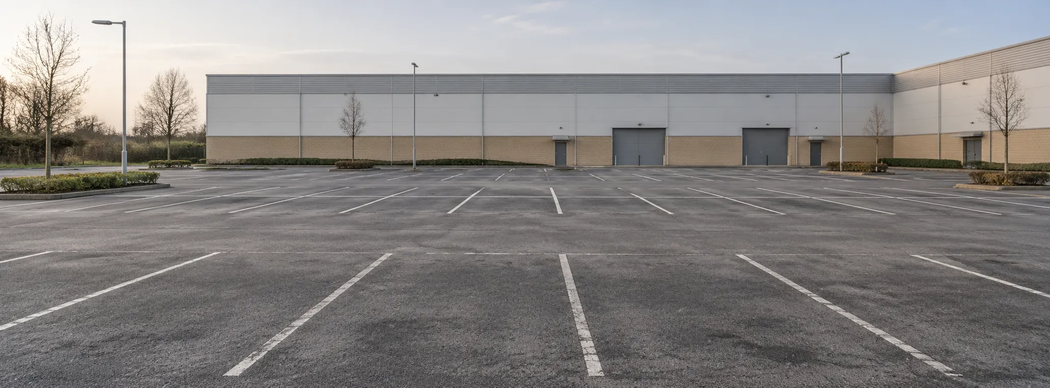Leerer asphaltierter Gewerbeparkplatz mit weißen Stellplatzmarkierungen, Laternen und Bäumen vor einer Halle