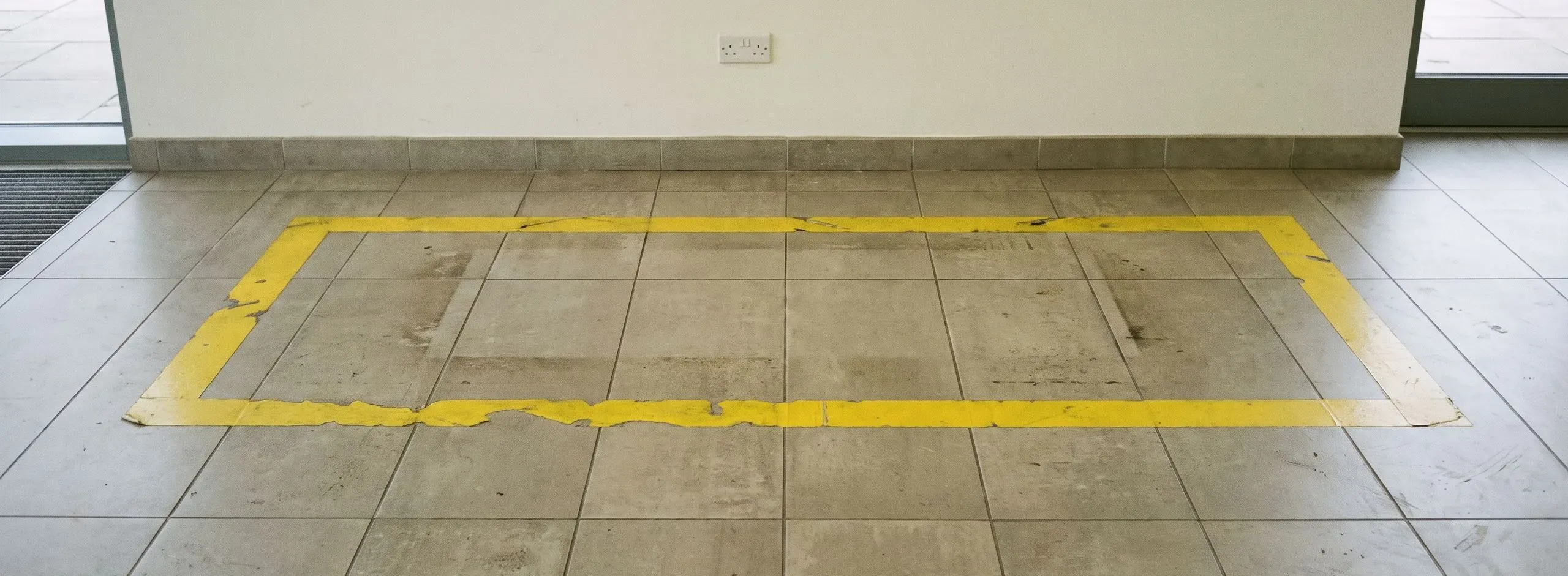 Leere gelbe Bodenmarkierung auf Fliesen, darüber eine Steckdose in der Wand